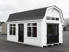 a small white building with black shingles on the roof and windows is in an empty lot