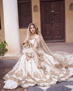 a woman in a white and gold wedding dress sitting on the ground with her veil draped over her head