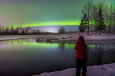 a person standing in front of a body of water under a green and purple aurora bore