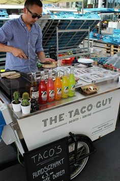 a man standing at a taco stand selling drinks and juices in front of him