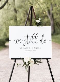 a welcome sign with white flowers on it and greenery in the background for an outdoor wedding