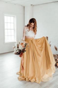 a woman is standing in a room wearing a long skirt and holding a flower bouquet