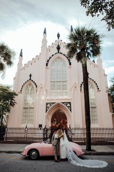 a bride and groom standing next to a pink car in front of an old church