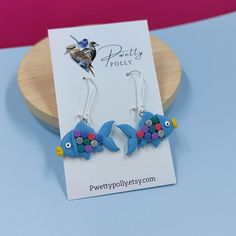 a pair of blue fish earrings with colorful beads on them sitting on a wooden stand