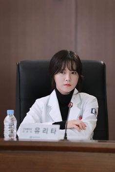 a woman sitting at a desk with a bottle of water in front of her and wearing a white lab coat