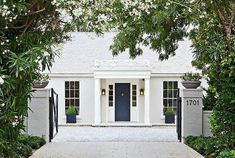 a white house with blue door and windows surrounded by greenery on either side of the entrance