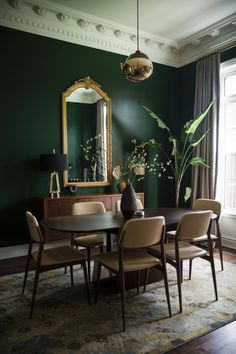 a dining room with dark green walls and wooden table surrounded by beige chairs, gold framed mirror and potted plant