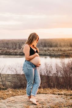 a pregnant woman standing on top of a hill next to a body of water at sunset