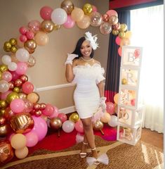 a woman standing in front of a balloon arch with balloons all around her and wearing a white dress