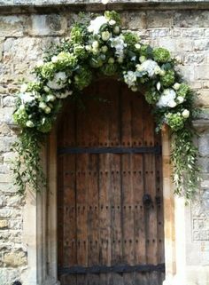 a wooden door with flowers and greenery on the outside, in front of a stone wall