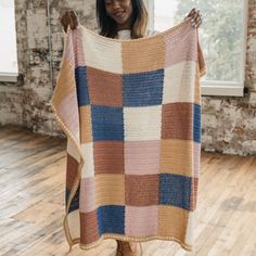 a woman holding up a multicolored blanket in front of a window with brick walls