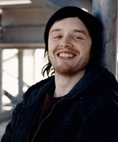 a man with long hair wearing a black hoodie smiles at the camera while leaning against a pole