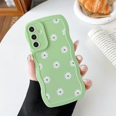 a hand holding a green case with white daisies on it and an orange croissant in the background