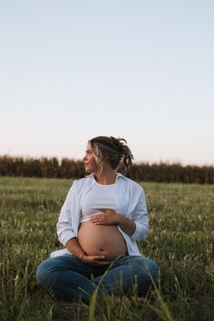 a pregnant woman sitting in the grass with her hands on her stomach and looking up at the sky