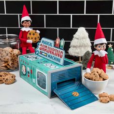 two elfs are standing next to a cookie machine and some cookies in front of them