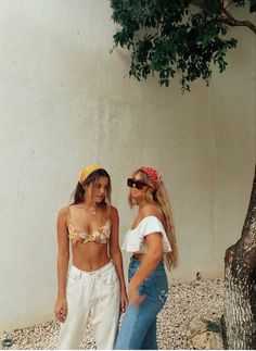 two women standing next to each other in front of a white wall and a tree