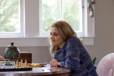 two women sitting at a table with chess pieces on the table and one woman standing next to her