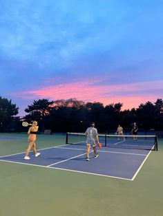 several people playing tennis on a blue and green court with trees in the background at sunset