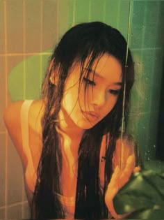 a woman with long black hair standing in front of a green tiled wall and holding onto a shower head