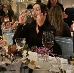 a woman sitting at a table drinking from a wine glass while surrounded by other people