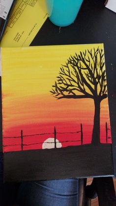 a person is holding up a painting with a sunset in the background and a tree painted on it