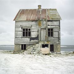 an old run down house sitting in the middle of some snow covered ground next to the ocean