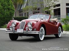 an old red car is parked in front of a building with trees and bushes behind it