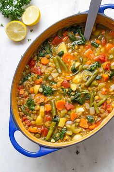 a blue pot filled with vegetable soup on top of a white counter next to lemons and parsley