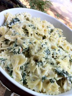 a white bowl filled with pasta and spinach on top of a wooden table next to a tree