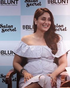 a woman sitting in a chair with her legs crossed, smiling at the camera and wearing an off shoulder dress