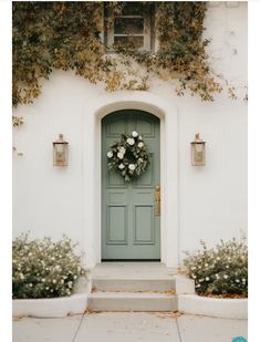 a green door with white flowers and greenery on the side of a house that has two planters in front of it