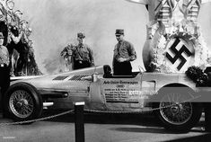 an old race car with men in uniform standing around it and another man looking on