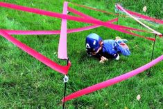 a small child in blue shirt and helmet crawling through pink tape on grass with red poles