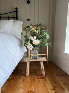 a vase with flowers sitting on a small wooden stool in front of a white bed