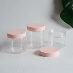 three glass jars with pink lids are sitting on a table next to a green plant