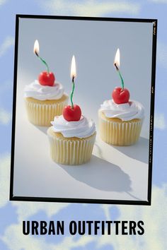 three cupcakes with white frosting and two cherries on top, one has a candle in the middle