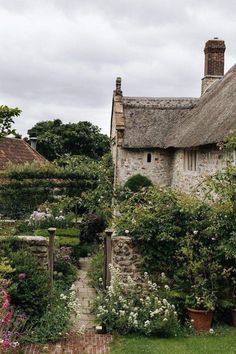 an old thatched roof house surrounded by greenery and flowers on a cloudy day