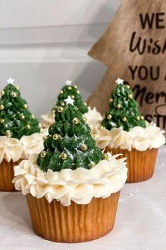 three cupcakes with white frosting and green trees on top are sitting next to a wooden sign