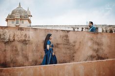 a man and woman standing next to each other on top of a cement wall with a building in the background