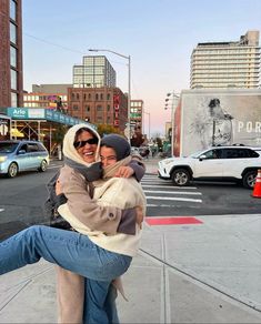 two people hugging each other on the sidewalk in front of some buildings and cars parked