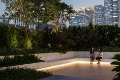 two women walking down a wooden walkway in front of tall buildings and trees at night