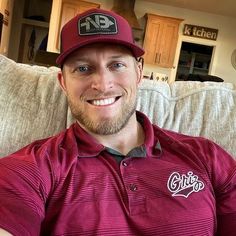 a man in a red shirt and hat sitting on a couch smiling at the camera