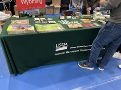 a man standing next to a green table covered in books and pamphlets at a convention