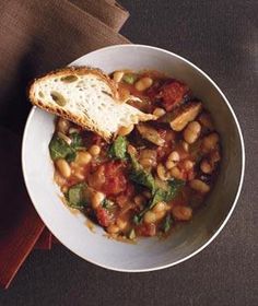 a white bowl filled with beans and vegetables next to a piece of bread on top of a table