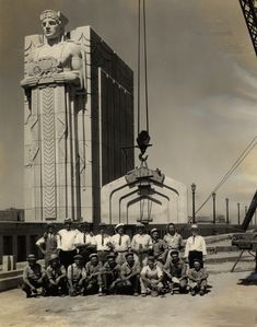 a group of men sitting next to each other in front of a tall building with a statue on top