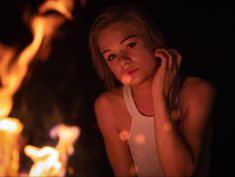 a woman is standing in front of a fire with her hand on her ear and looking at the camera