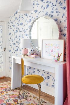 a white desk with a mirror and yellow chair in front of flowers on the wall