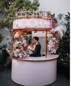 a bride and groom kissing in front of a booth with flowers on the wall behind them