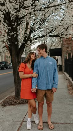 a man and woman standing next to each other on the sidewalk in front of a tree