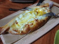 a piece of bread with cheese on it and a fork stuck into it, sitting on a cutting board